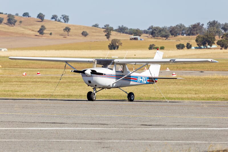 A white Cessna 150G with blue trim parked on an airfield apron in front of dry hills, its high wing braced by a single strut.