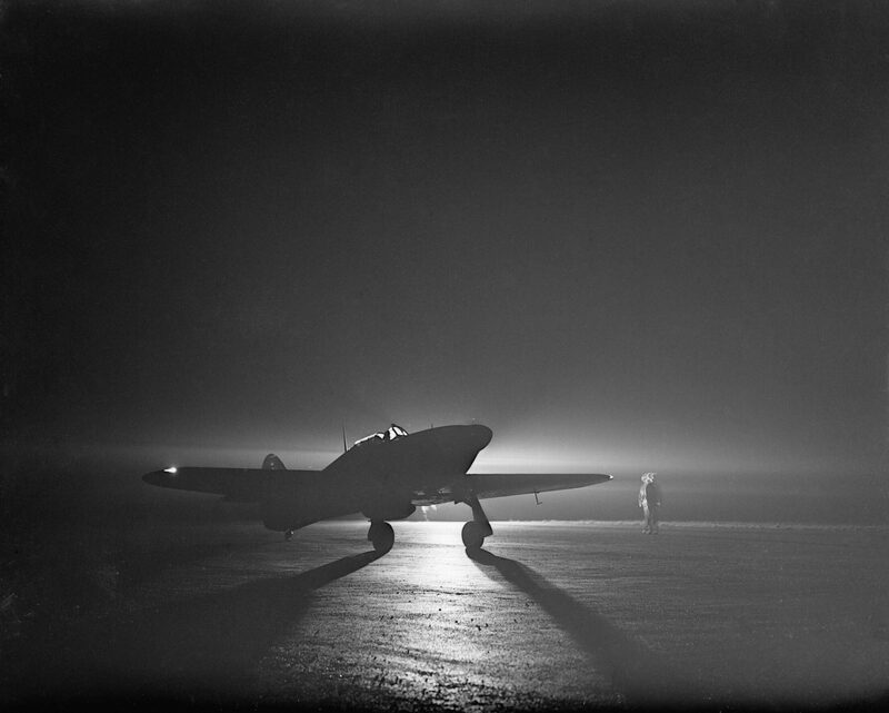 A Hawker Hurricane Mk I of No. 85 Squadron taxiing in darkness by the light of a flare at Debden in March 1941, its silhouette lit from behind.